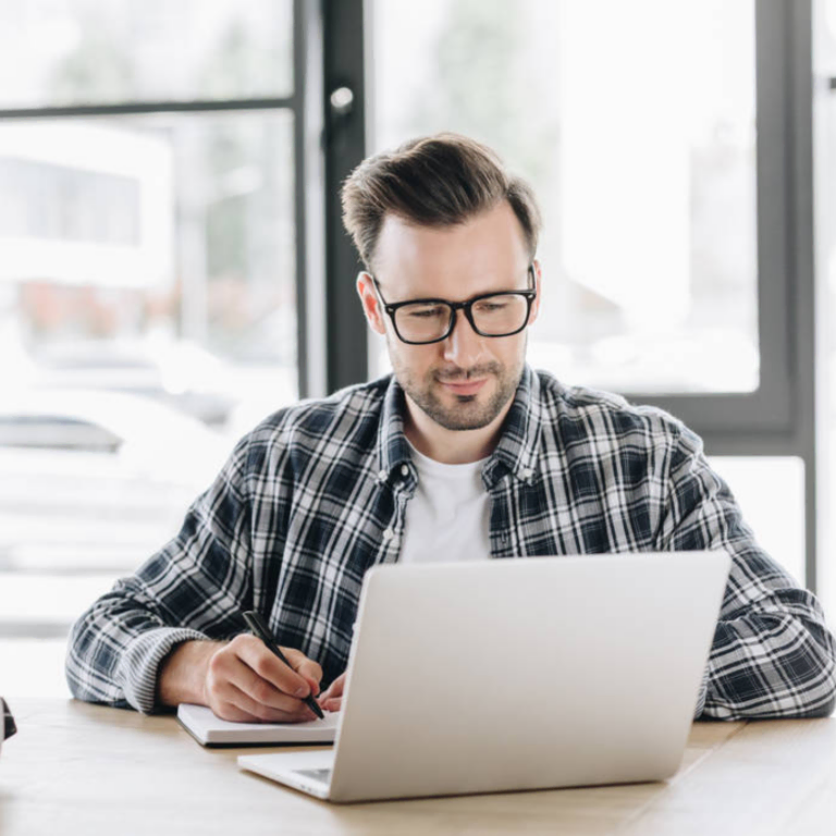 Man in glasses writing notes beside a laptop