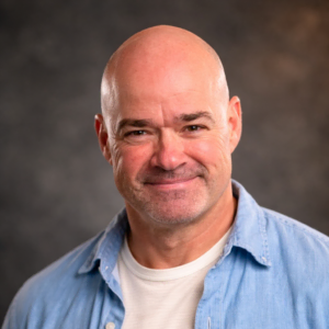 Professional headshot of a smiling bald man in a denim shirt