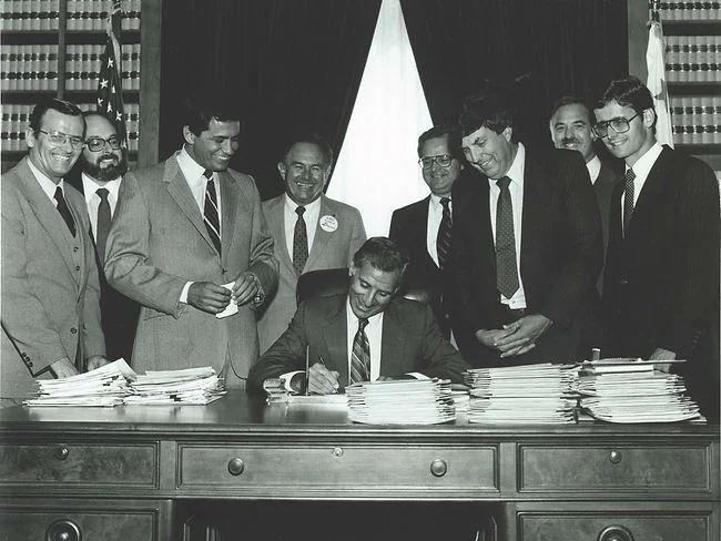 Business professionals gathered around a desk during a signing event in a formal setting.