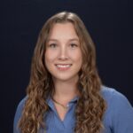 Smiling young woman with long curly hair wearing a blue shirt against a dark background.