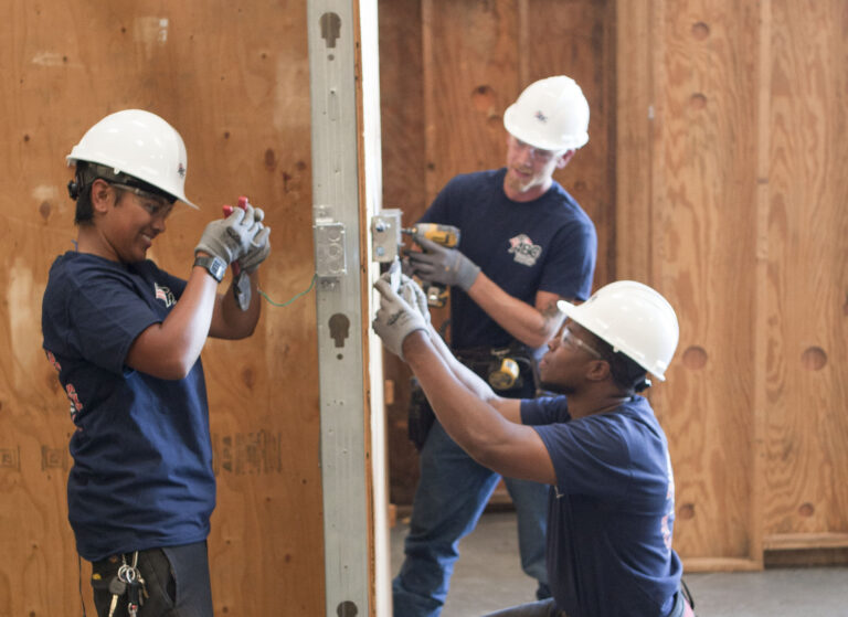 Three workers wearing hard hats assemble a wall panel in a construction site.